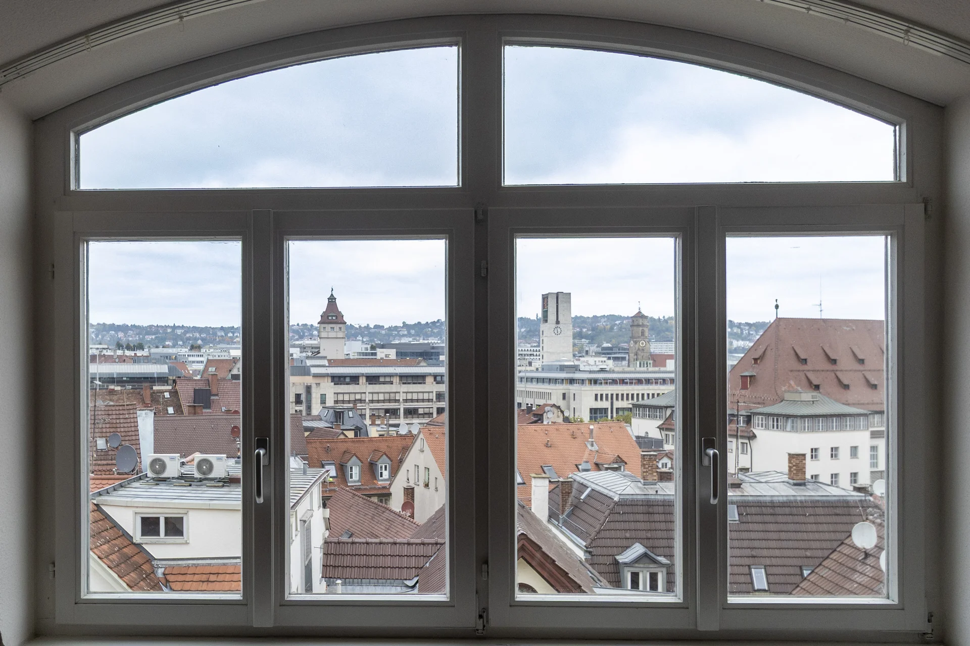 Blick durch Bogenfenster auf Stadtpanorama mit roten Ziegeldächern, Kirchtürmen und modernen Gebäuden bei bewölktem Himmel.