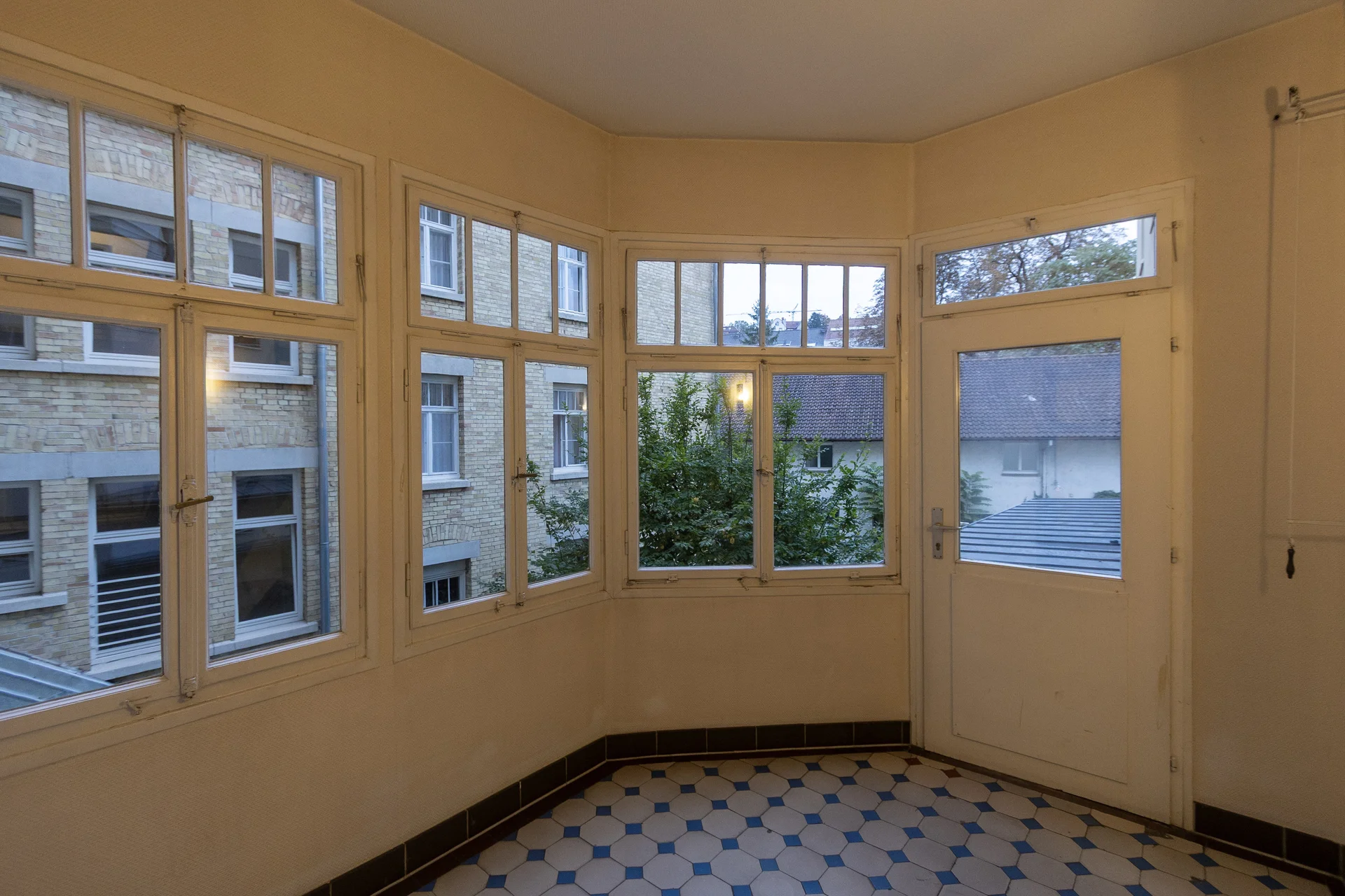 Heller Raum mit großen Fenstern, gelben Wänden und schwarz-weiß-blauem Fliesenboden, Blick auf Backsteingebäude