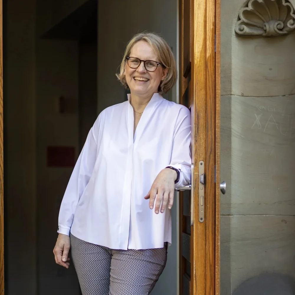 Frau mit Brille und blonder Kurzhaarfrisur in helllila Bluse steht lächelnd in steinerner Türöffnung mit Holzrahmen.